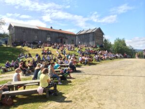 Gammel gård med mange mennesker som sitter på bakken og lytter til musikk.