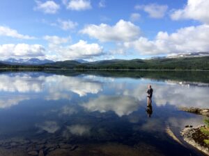 Mann står uti vatnet og fisker med fluestang, blankstille vatn og fjell rundt.