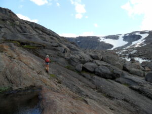 Mann og dame går opp en bergskråning. Blå himmel og snø i fjellene bak.