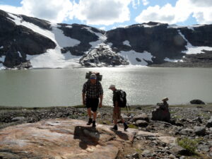To menn på tur i fjellet, med fjell, snø og et vatn i bakgrunnen.